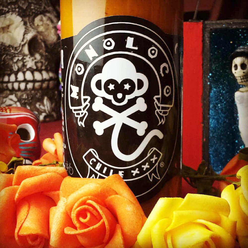 A close-up view of a MONOLOCO Chile XXX Hot Sauce bottle against a festive backdrop featuring Mexican Day of the Dead skull figurines and colorful flowers, showcasing the sauce's vibrant cultural presentation.