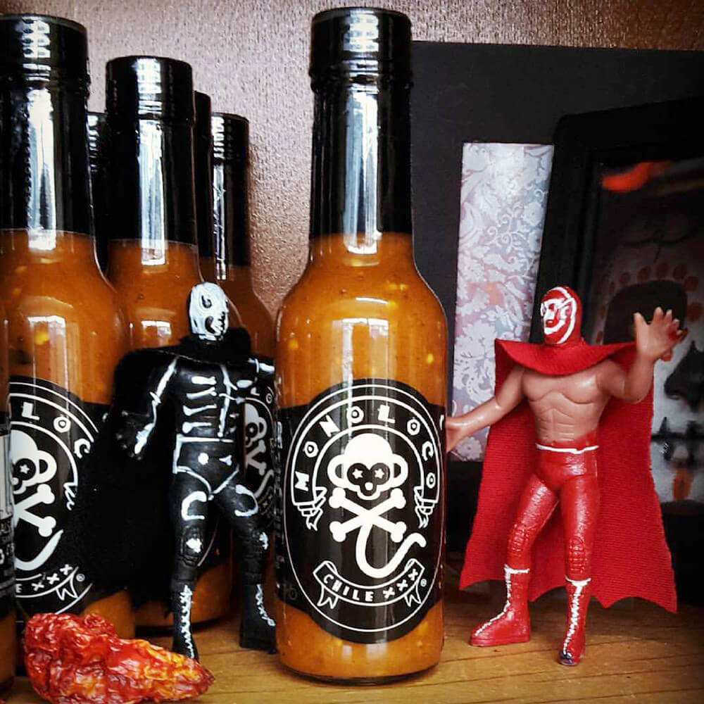 A collection of MONOLOCO Chile XXX Hot Sauce bottles displayed with vibrant Mexican wrestling figurines, highlighting the cultural and playful presentation of this medium-heat sauce.