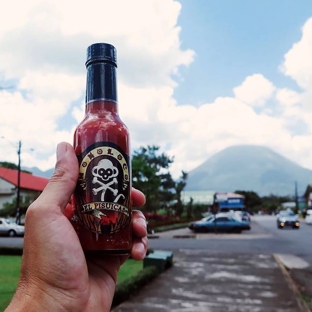 Hand holding a bottle of MONOLOCO El Pisuicas Hot Sauce with a volcano in Costa Rica in the background, showcasing the sauce's cultural and geographical roots.