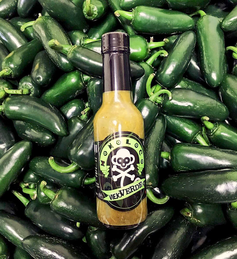 MONOLOCO Viaje Verde Hot Sauce bottle surrounded by fresh Jalapeño peppers, highlighting the sauce's fresh and herb-infused qualities.